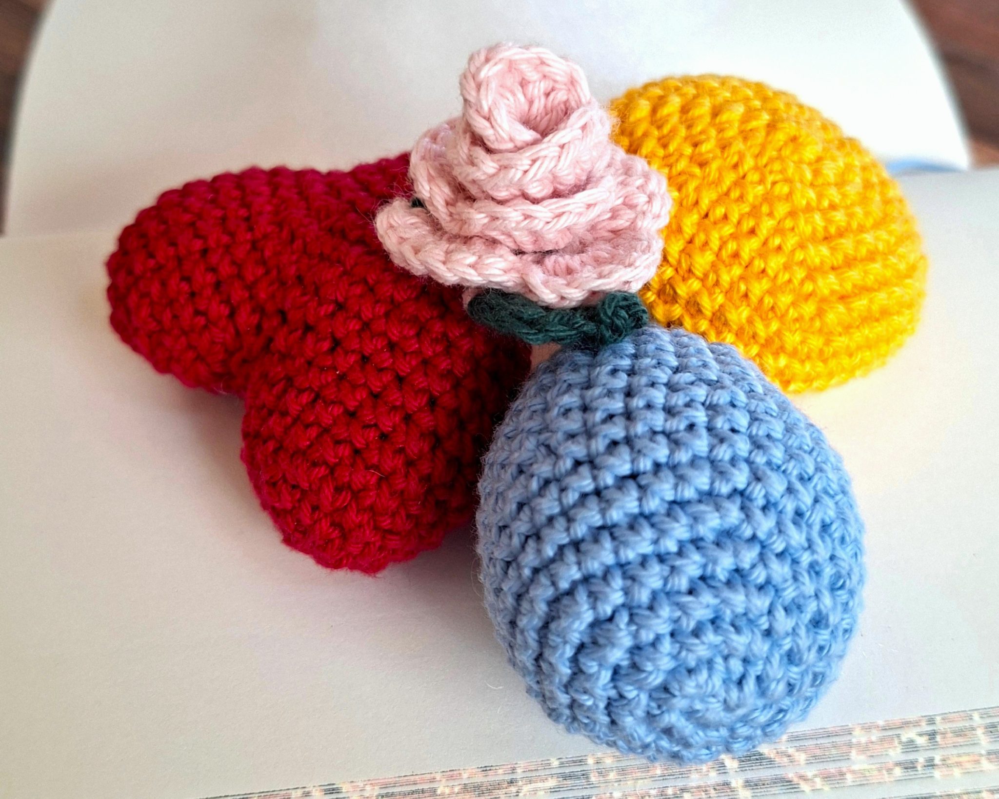 Trois formes crochetées en rouge, jaune et bleu, représentant un cœur, une goutte et une demi-sphère, avec une rose pastel au centre. Amigurumis en devenir.