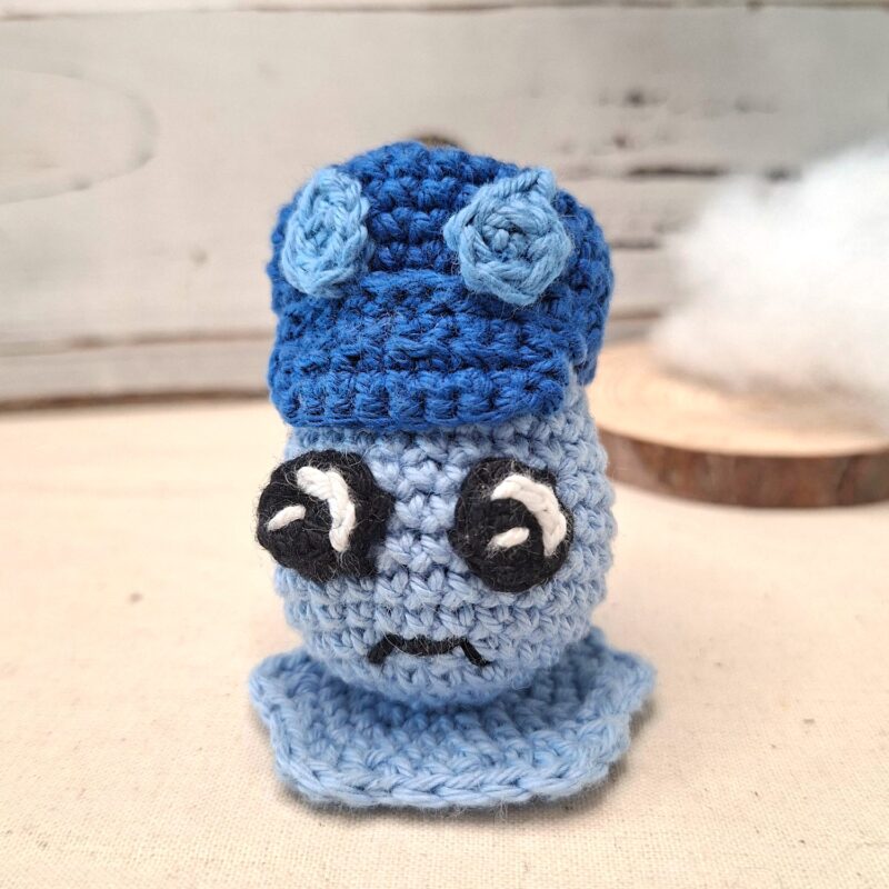 Amigurumi en forme de goutte bleue au crochet, portant une casquette sombre, photographié de face.