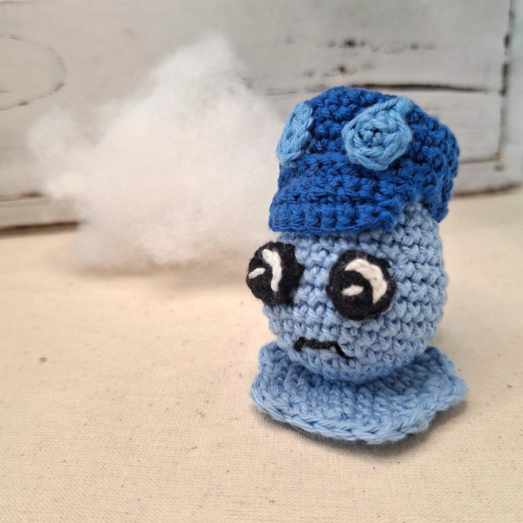 Amigurumi crochet bleu clair en forme de goutte, avec casquette sombre, posé devant un nuage blanc, symbolisant la tristesse retenue.