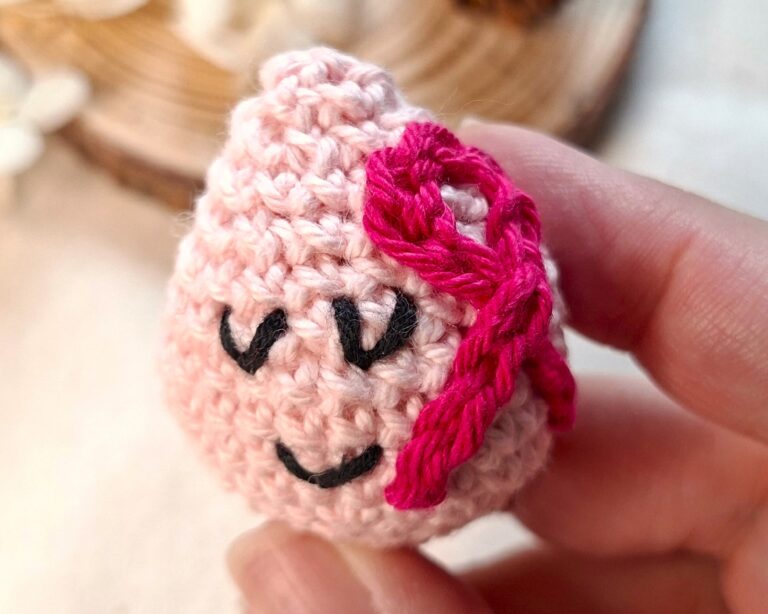 Goutte rose au crochet tenue entre les doigts, symbole poétique d’Octobre Rose et de solidarité féminine – création Clairimagine.