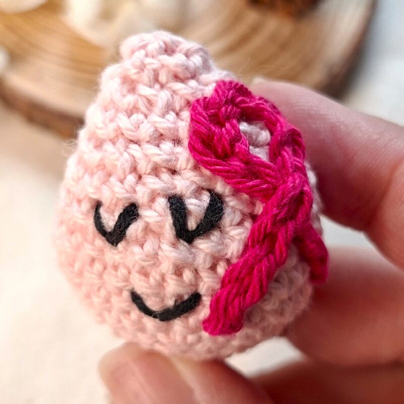 Goutte rose au crochet tenue entre les doigts, symbole poétique d’Octobre Rose et de solidarité féminine – création Clairimagine.