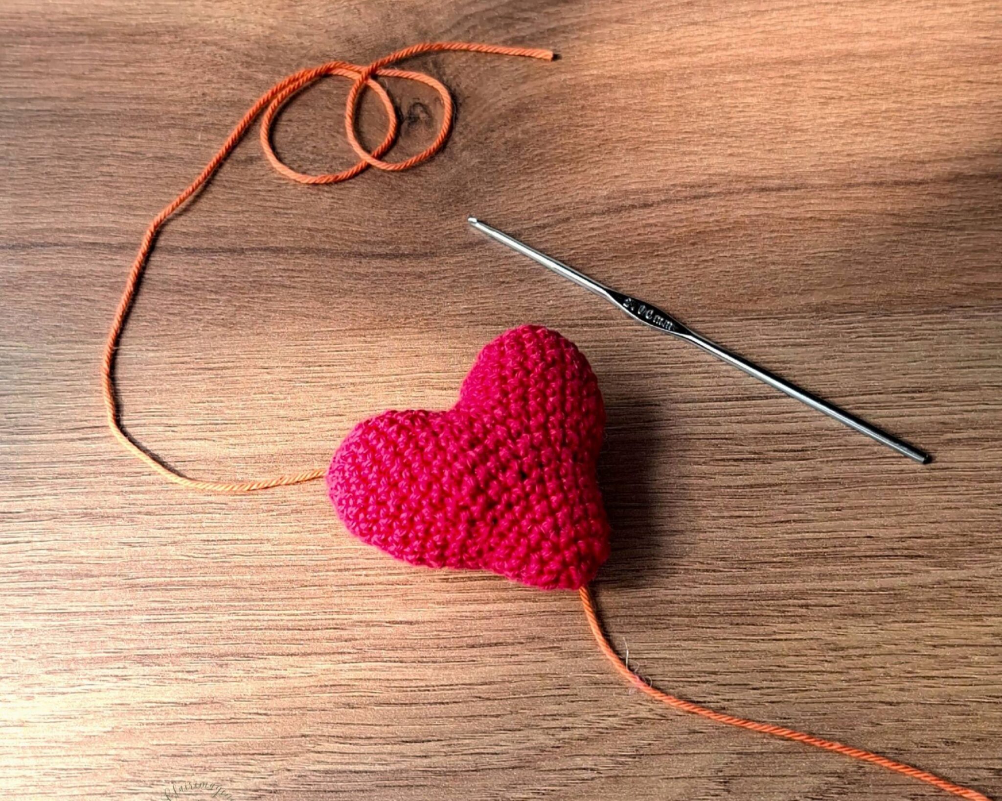 Cœur au crochet rouge posé sur une table en bois, relié à un fil orange et à un crochet métallique, symbole de création et de patience artisanale.