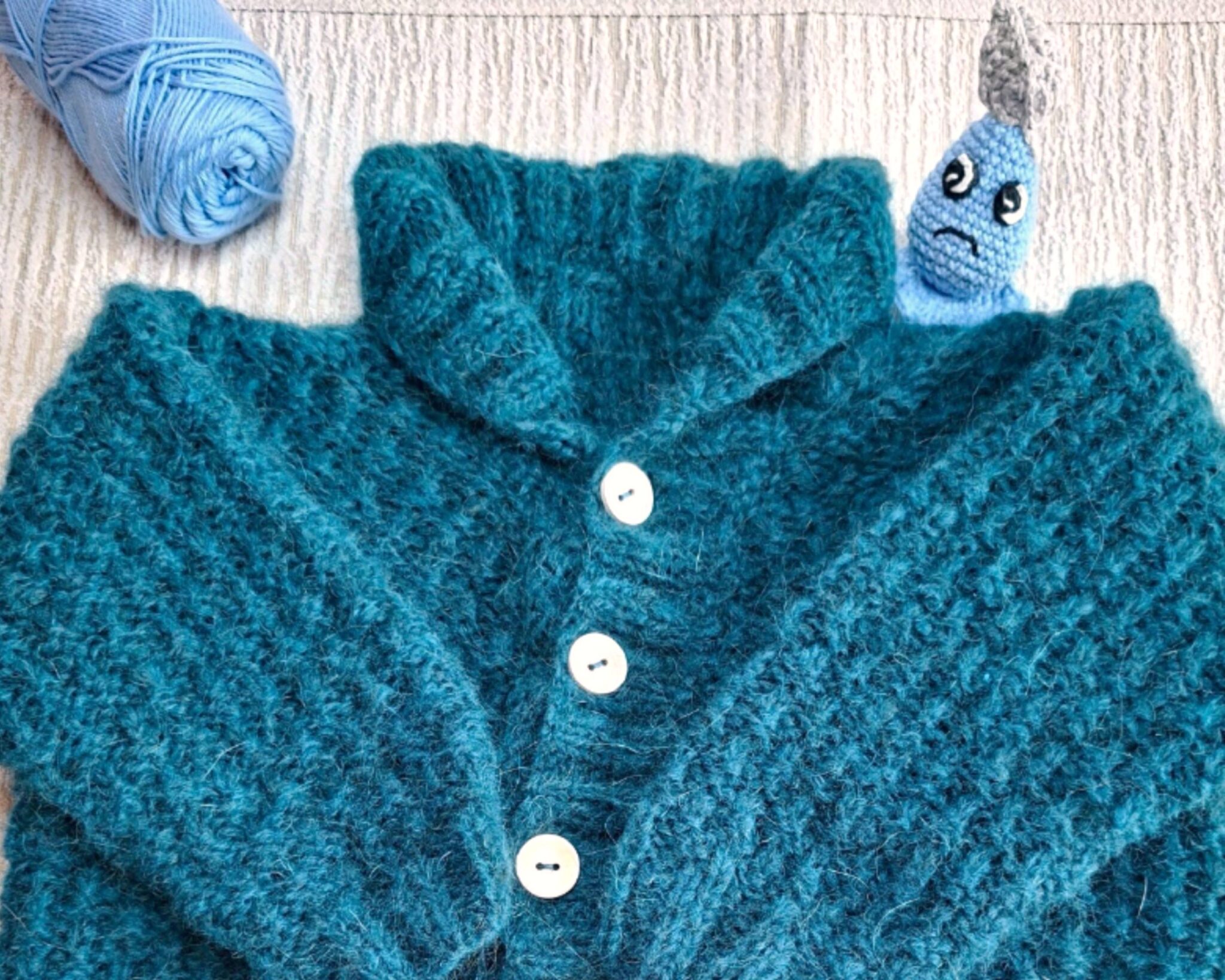 Gilet en laine tricoté main, couleur bleu canard, accompagné de Plumélia, la petite goutte de douceur. Une image poétique signée Clairimagine, symbole de chaleur et de tendresse hivernale.
