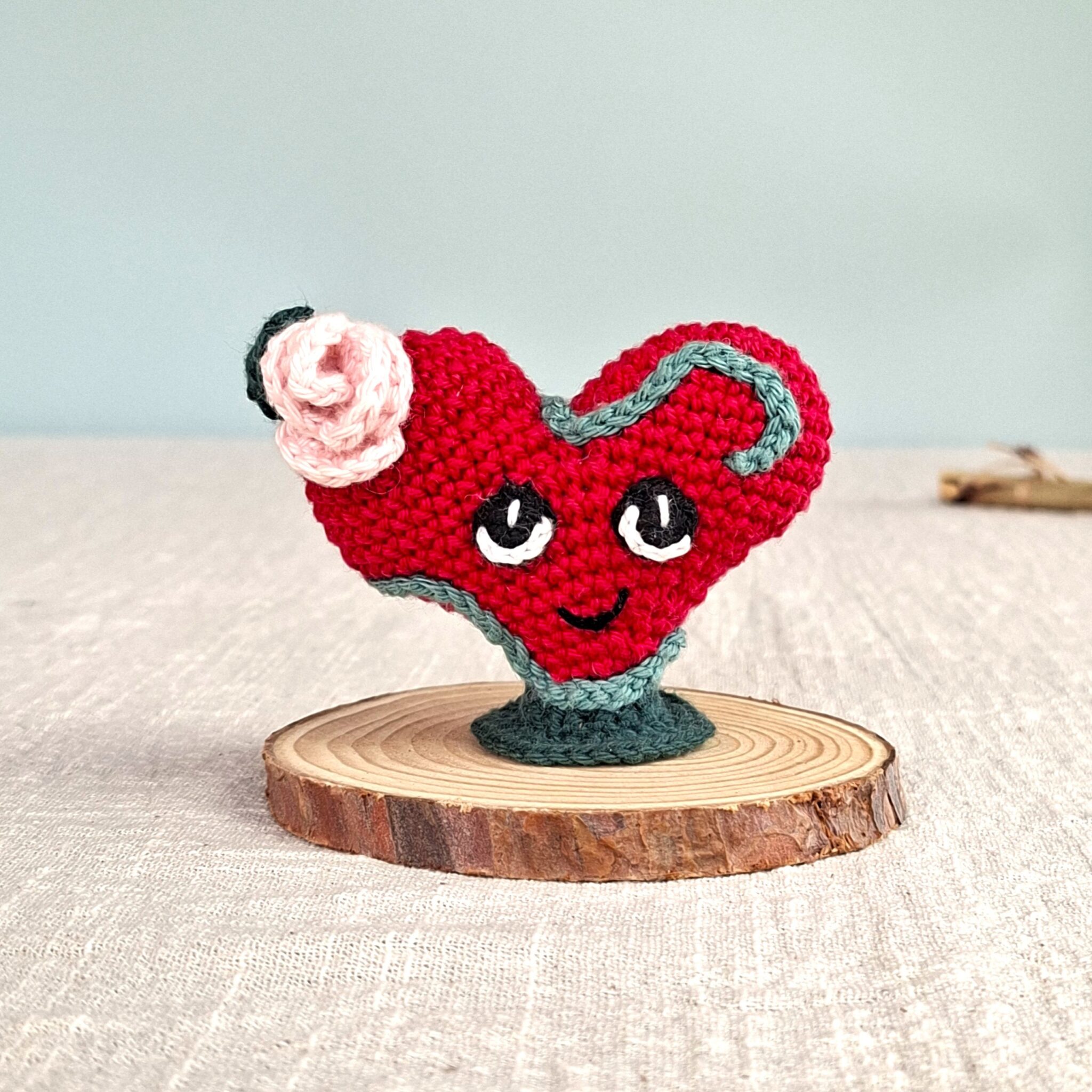 Amigurumi cœur rouge Lianis avec liane verte et rose crochetée, symbole d’amour passionné, posé sur socle en bois