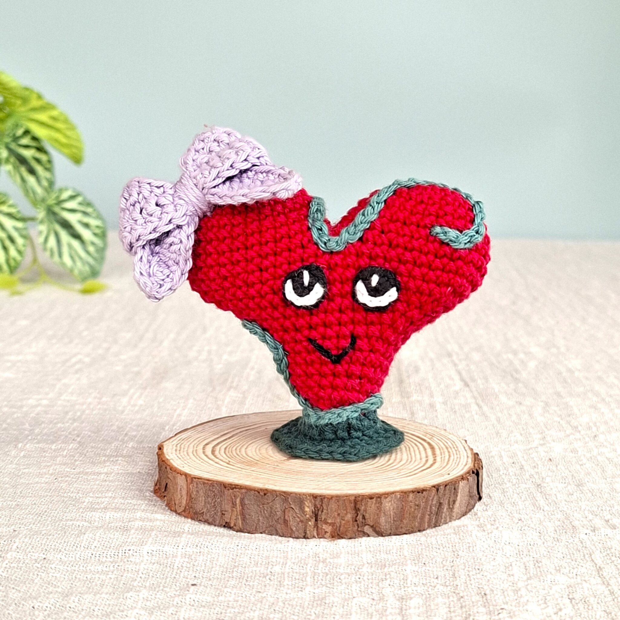 Amigurumi cœur rouge Lianis avec liane verte et nœud lavande, symbole d’amitié et complicité, posé sur socle en bois