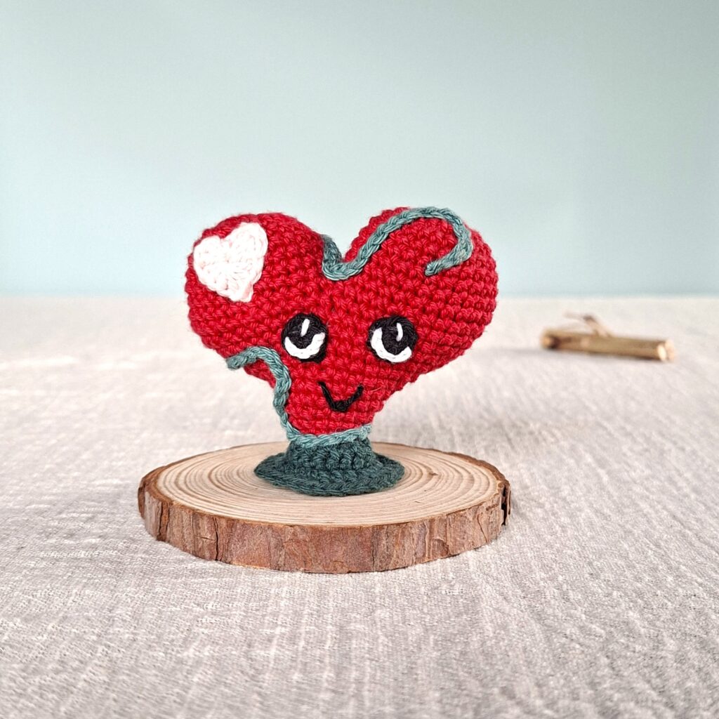 Amigurumi cœur rouge Lianis avec liane verte, symbole d’amour inconditionnel, posé sur socle en bois