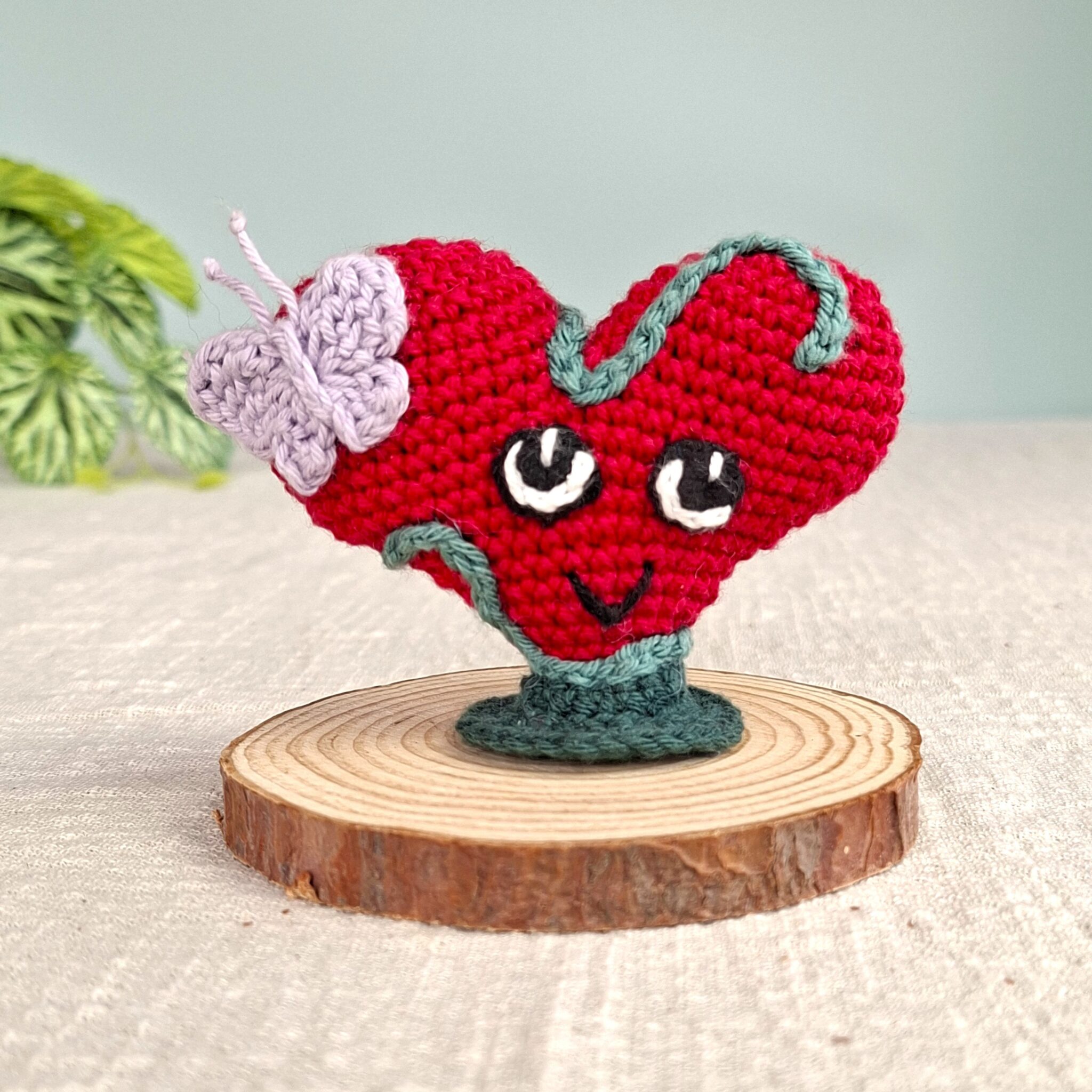 Amigurumi cœur rouge Lianis avec liane verte et papillon lavande, symbole d’amour universel, posé sur socle en bois