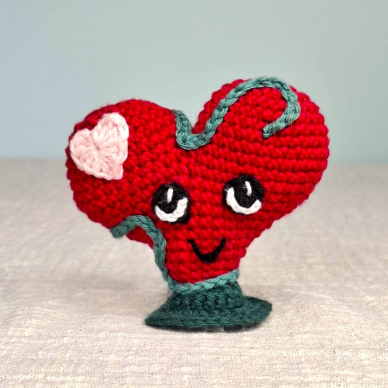 Amigurumi Lianis, cœur rouge au crochet avec liane verte et petit cœur rose, sur socle vert, vue de face